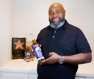 Glenn Thomas, Founder of John Lee Hooker Legacy Spirits, holding a bottle of award winning Boogie Chillen Bourbon. In the background, a framed Hollywood Walk of fame star and multiple bottles of the bourbon are displayed. The bottle's label features an im