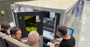 People examining a newly cut metal component from a laser cutting machine in a busy workshop environment.