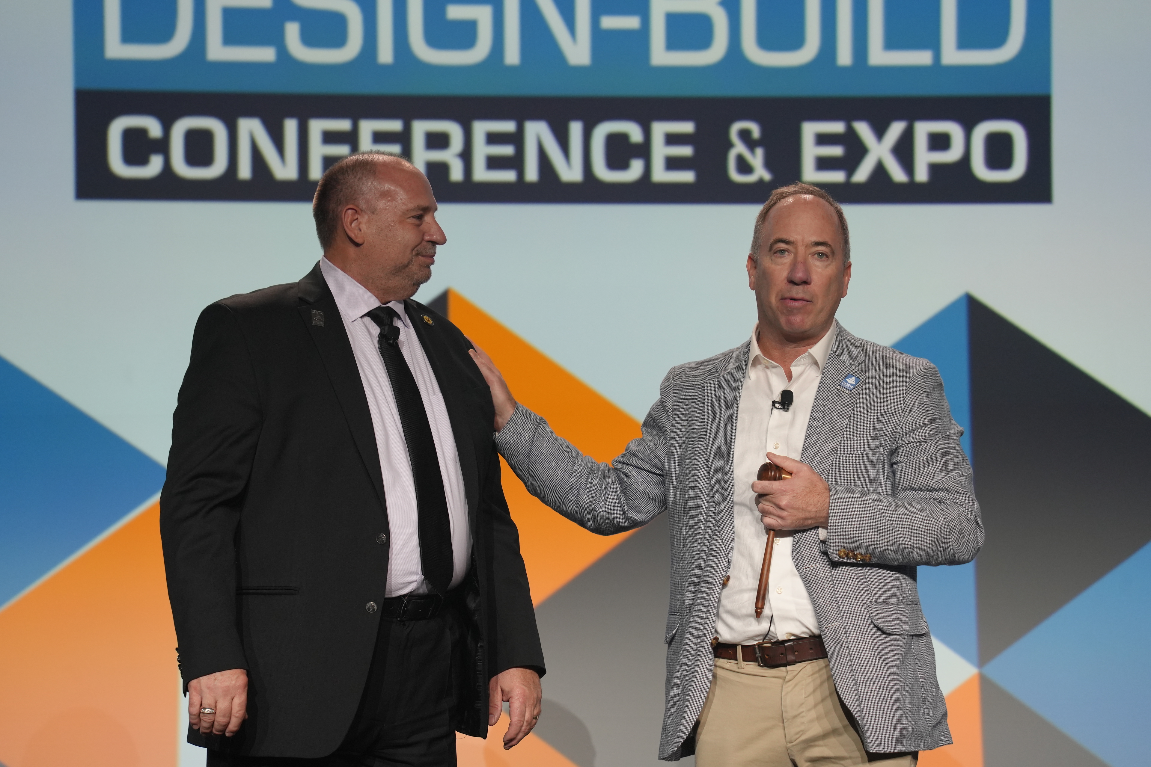 Two speakers on stage at the Design-Build Conference & Expo, standing in front of a backdrop with the event’s logo. One is wearing a dark suit and tie, while the other wears a light gray blazer and holds a gavel, gesturing as he speaks