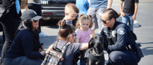 A black dog being petted by people with a person in a police uniform kneeling beside it.