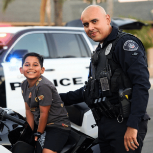 A police officer standing next to another individual seated on a motorcycle, with a patrol car in the background.