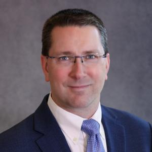 Small in dark suit and tie with glasses smiling and directly looking at the camera