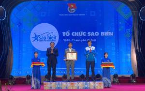  Award ceremony by Sao Biển - Room for Education and Ho Chi Minh Communist Youth Union in Hanoi, 2016. Central person holds a framed certificate, flanked by two others with certificates. Two women in traditional attire hold red and gold trays. Colorful stage backdrop.