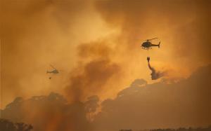 A helicopter flies over some hills, dropping water over the fire below.