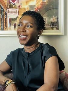 A professional headshot of Moniek James, a smiling woman with short, natural hair, wearing a sleek black blouse and accessorized with bracelets, seated against a backdrop of a framed cityscape painting.