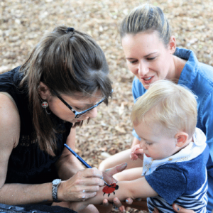 Adult painting on a child's arm outdoors while another adult watches.