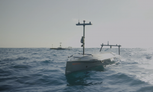 A gray autonomous boat approaches the camera with multiple other boats in the background.