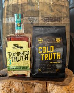 A bottle of Tarnished Truth Whiskey alongside a bag of Cold Truth Whiskey Barrel-Aged Coffee are shown sitting in front of a barrel.