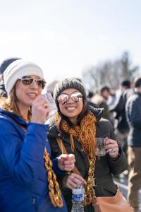 Two women in winter gear are holding beer sample glasses and wearing necklaces made from pretzels at Kennett Winterfest.