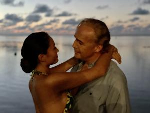 Still photo from Waltzing with Brando featuring Heiae (Heather) Touniou as Tarita and Billy Zane as Marlon Brando. Touniou, in a tropical-print bikini, embraces Zane, who wears a light shirt. They share an intimate moment against a serene ocean sunset.