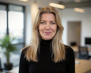 A professional portrait of Ela Slutski, Managing Director of Cirrus Assessment. She has long blonde hair and is wearing a black turtleneck sweater. She is standing in a modern office setting with large windows, natural light, and an open workspace in the 