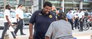 A community safety officer engages with a citizen amidst a bustling urban scene with police officers and pedestrians in the background.