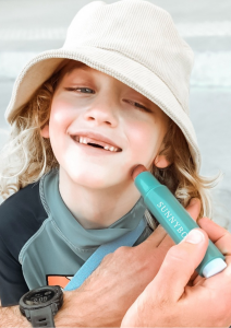 A happy blonde child with a bright smile has sunscreen applied with a SUNNYBOD Refillable Sunscreen Brush, promoting healthy sun safety habits.