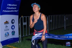 A woman in a black tank top, running tights, and a colorful cap crosses the finish line at night, breaking the race tape with her hand. She clenches her fist and laughs with joy, filled with emotion after completing the third race of the World Marathon Ch