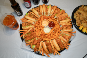 A mouthwatering po’ boy platter loaded with crispy, golden-fried seafood nestled in fresh baguettes with lettuce. The sandwiches are arranged around a generous serving of seasoned fries, accompanied by dipping sauces including ketchup, a creamy white sauc