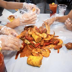 A flavorful seafood boil piled high with fresh shrimp, crawfish, crab legs, sausage, corn on the cob, and potatoes, all drenched in a rich, buttery spicy sauce. Diners, wearing plastic gloves, dive into the feast, peeling shrimp and cracking crab legs for