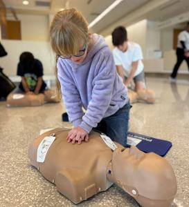 A young girl in a purple hoodie performs CPR on a mannequin during National Youth Heart Screening Day. Other youth in the background also practice CPR, learning critical life-saving skills to respond to sudden cardiac arrest.