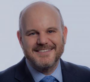 "Professional headshot of a smiling man wearing a navy suit jacket, light blue dress shirt, and blue patterned tie. He has a bald head and a full beard, and is photographed against a light gray background."