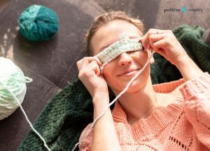 a woman in a peach colored crochet sweater is smiling and laying on the floor with a peace of crochet fabric on her eyes
