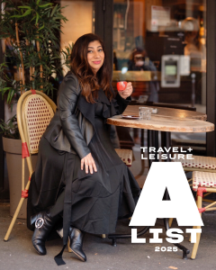 A woman with long, wavy brown hair and a warm smile sits at an outdoor café table, holding a small red espresso cup. She is dressed elegantly in an all-black ensemble, featuring a flowing skirt, a stylish leather jacket, and heeled boots adorned with a de