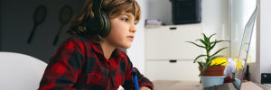 A young child with shoulder-length brown hair, wearing headphones and a red plaid shirt, sits at a desk using a laptop. The background features a white cabinet, a potted plant, and a well-lit room with tennis rackets hanging on the wall.