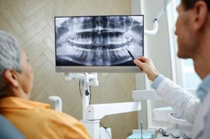 A side view close-up of a male dentist pointing at a teeth X-ray image on a screen while discussing results with a senior female patient.