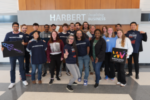 A diverse group of young entrepreneurs and mentors pose enthusiastically in front of the Harbert College of Business sign. They are wearing event-branded t-shirts that read "Build a Business in a Weekend" and holding signs with startup-themed graphics, in