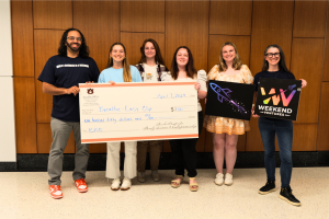 A group of six smiling individuals stand in front of a wooden-paneled wall, holding an oversized check for $150 awarded to "Breathe-Easy Clip" from Auburn University’s Harbert College of Business. Two members hold startup-themed signs, including a rocket