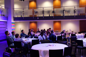 A group of attendees sits at round tables in a dimly lit event space with a modern design, attentively listening to a speaker. The room is illuminated with blue and purple lighting, and a "Weekend Ventures" banner is visible in the background. Some attend