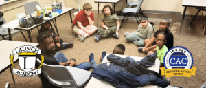 A group of children sits in a circle on the floor of a classroom, engaged in a discussion, with educational materials nearby.