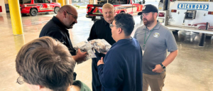 A group of people stands inside a fire station, discussing and exchanging items with emergency vehicles in the background.