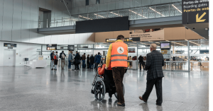 An airport terminal with passengers moving towards boarding gates. A staff member wearing an orange high-visibility vest with a wheelchair accessibility symbol is assisting a passenger in a wheelchair. Another elderly man walks alongside them. The termina