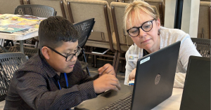 A boy in a long-sleeved black shirt works on a computer with a woman in glasses sitting at right.