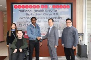 A group of five people pose in front of a welcome sign. Two men in the center, Dr. Raphael Olaiya and Dr. Koji Kurusu, shake hands, while others stand or sit nearby, smiling.