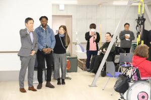 A man in a suit gestures while speaking, as others, including a man in a wheelchair, observe in a rehabilitation facility.