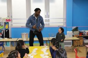 A man playfully interacts with a laughing child in a classroom while other children sit at a table with bowls of food.