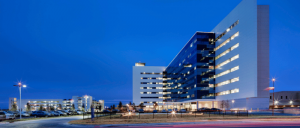 Modern hospital building illuminated at dusk, showcasing large windows and a sleek design, surrounded by a parking area.