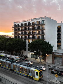 Liv DTLA, a six-story residential building with balconies, stands along a busy street in Downtown Los Angeles at sunset. A Metro light rail train passes in the foreground, and the warm glow of the setting sun reflects off the building’s white facade. Lush