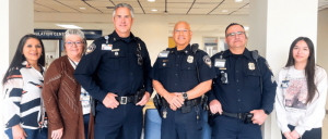 A group of four police officers in dark uniforms stands proudly with three individuals in casual clothing, posed together in a well-lit hallway.