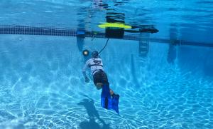 A person is underwater in a swimming pool inspecting a light as he is breathing from Nomad Mini with it floating above him on the surface.