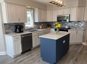 A modern, newly refaced kitchen featuring light wood grain cabinets, a navy blue island, and a sleek white quartz countertop. The backsplash consists of blue and gray mosaic tiles, complementing the stainless steel appliances. A glass pendant light hangs 