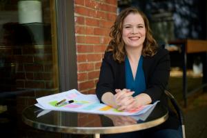 Sara is sitting outside at a glass table in front of a brick wall. The table has large sheets of paper on top with sticky notes all over it. Sara's hands are folded, resting on the table. Sara has shoulder length wavy dark blonde hair and is a 40-somethin