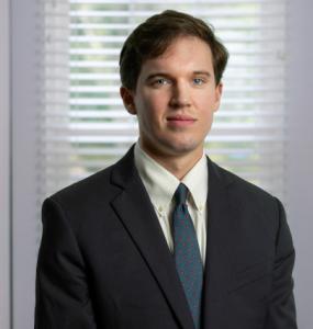 Professional headshot of Joseph T. Siegel from Carr Legal Group. He is wearing a dark suit jacket, white button-down shirt, and patterned tie. He has short brown hair and is standing in front of a window with white blinds, looking directly at the camera w