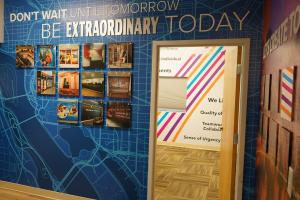 An interior wall at Heritage Signs & Displays' Arlington, VA office, featuring a blue wall wrap outlining DC Metro geography, motivational text presented through dimensional lettering, and acrylic-mounted photos showcasing past projects.
