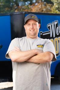 Brian Hensley, a middle-aged man with a beard and light skin stands confidently with his arms crossed, smiling slightly. He is wearing a gray T-shirt with a 'Weco Rooter' logo on the chest and a black baseball cap with the same branding. Behind him is a b