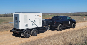 A white Flexx Mobile DC Fast Charger trailer is being towed by a black electric Hummer on a dirt road through rural Texas countryside, surrounded by open fields under a clear blue sky.