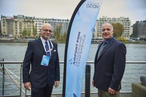 William L. Neece and Roberto Castiglioni MBE standing beside a banner for the Airport PRM Leadership Conference, with a river and buildings in the background.