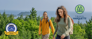 Two hikers navigate a scenic forest trail, surrounded by lush green trees and a coastal backdrop, promoting outdoor exploration.