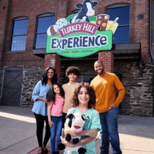A group of five people stands in front of the Turkey Hill Experience sign, holding a stuffed cow toy, in an outdoor setting.