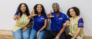 Four individuals sit together on a wooden bench, wearing colorful branded shirts and making a sign with their hands in a friendly pose.